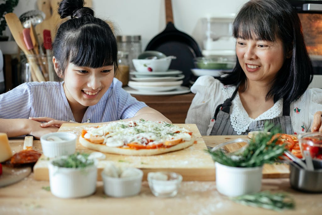 Satisfied Asian women different ages spending time together while cooking yummy pizza on cozy kitchen with various utensil and food around